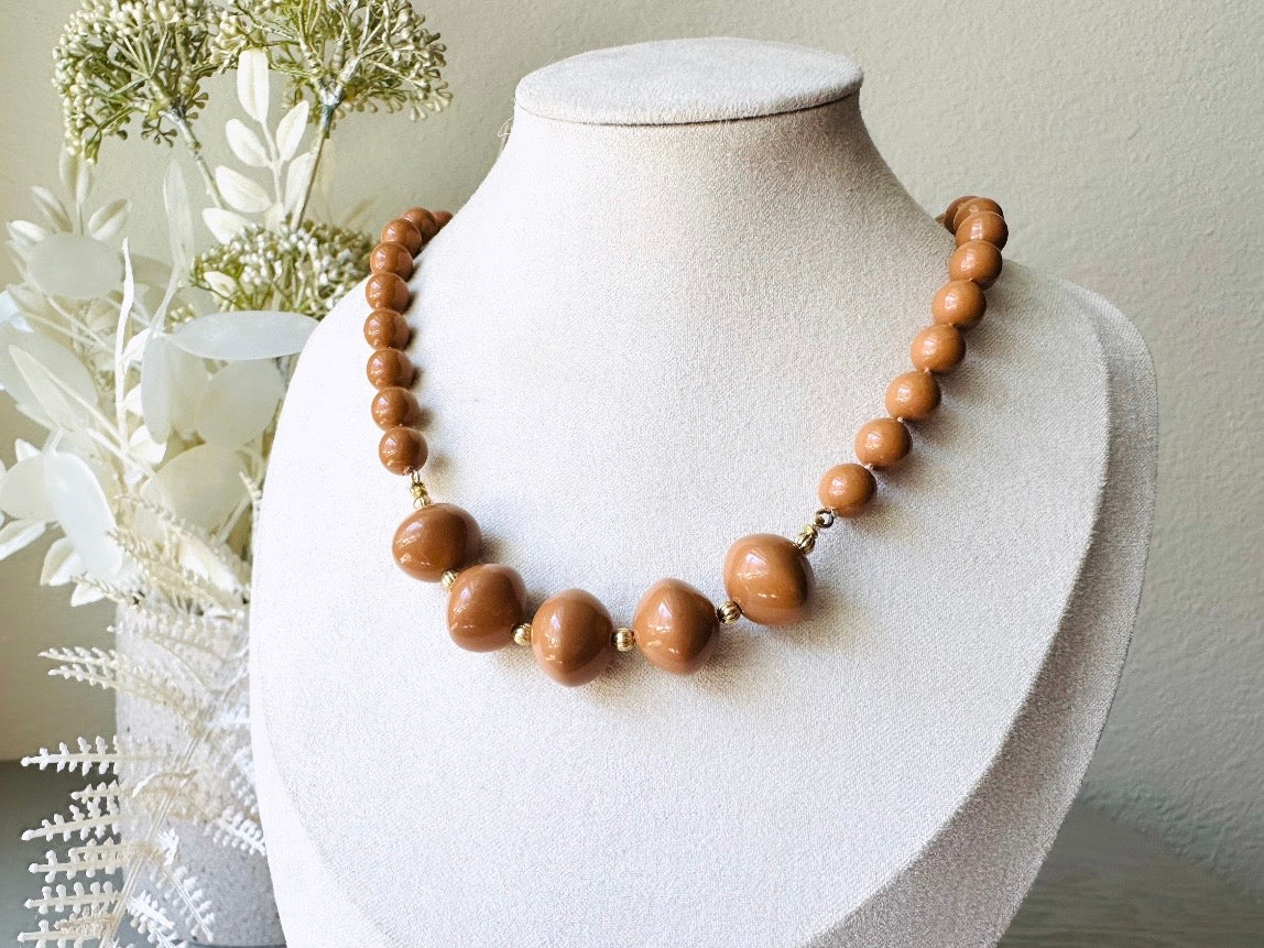 A vintage Park Lane designer necklace. The necklace is composed of 6mm caramel round beads with 5 bigger rondelle beads in the center with tiny gold spacer beads between. The necklace is on a bust display on a light gray wood table with a floral background.