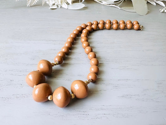 A vintage Park Lane designer necklace. The necklace is composed of 6mm caramel round beads with 5 bigger rondelle beads in the center with tiny gold spacer beads between. The necklace is on a light gray wood table with a floral background.
