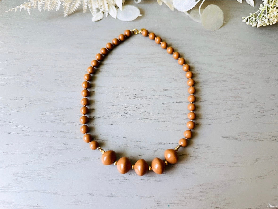 A vintage Park Lane designer necklace. The necklace is composed of 6mm caramel round beads with 5 bigger rondelle beads in the center with tiny gold spacer beads between. The necklace is on a light gray wood table with a floral background.