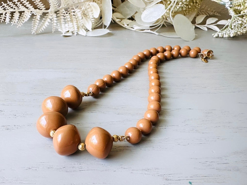 A vintage Park Lane designer necklace. The necklace is composed of 6mm caramel round beads with 5 bigger rondelle beads in the center with tiny gold spacer beads between. The necklace is on a light gray wood table with a floral background.