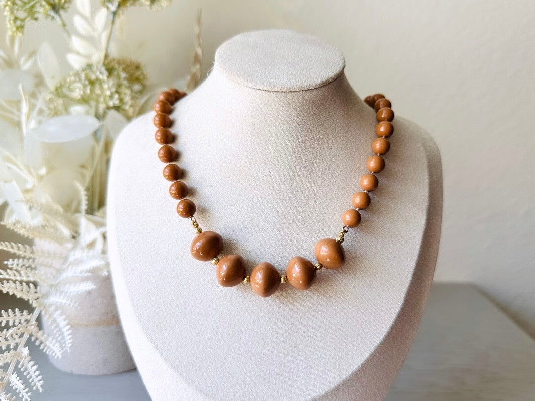 A vintage Park Lane designer necklace. The necklace is composed of 6mm caramel round beads with 5 bigger rondelle beads in the center with tiny gold spacer beads between. The necklace is on a bust display on a light gray wood table with a floral background.