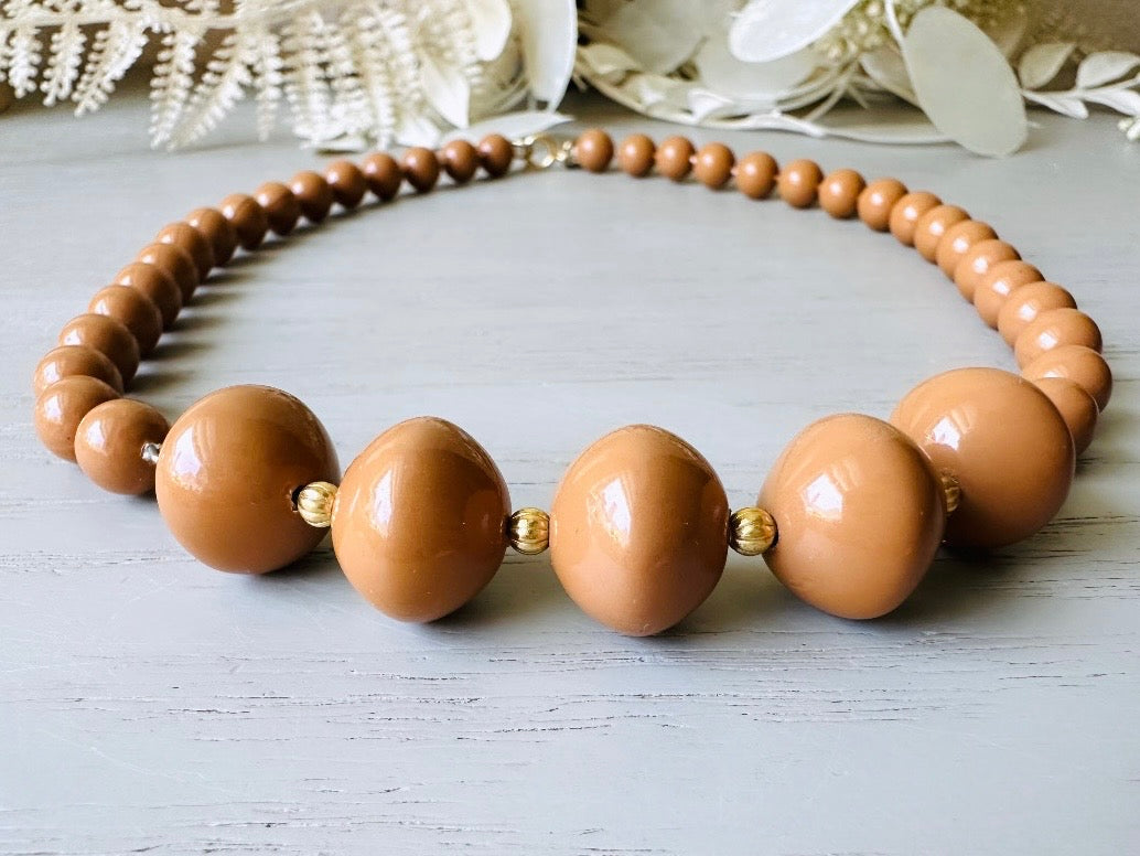 A vintage Park Lane designer necklace. The necklace is composed of 6mm caramel round beads with 5 bigger rondelle beads in the center with tiny gold spacer beads between. The necklace is  on a light gray wood table with a floral background.