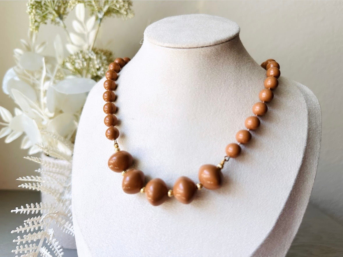 A vintage Park Lane designer necklace. The necklace is composed of 6mm caramel round beads with 5 bigger rondelle beads in the center with tiny gold spacer beads between. The necklace is on a bust display on a light gray wood table with a floral background.