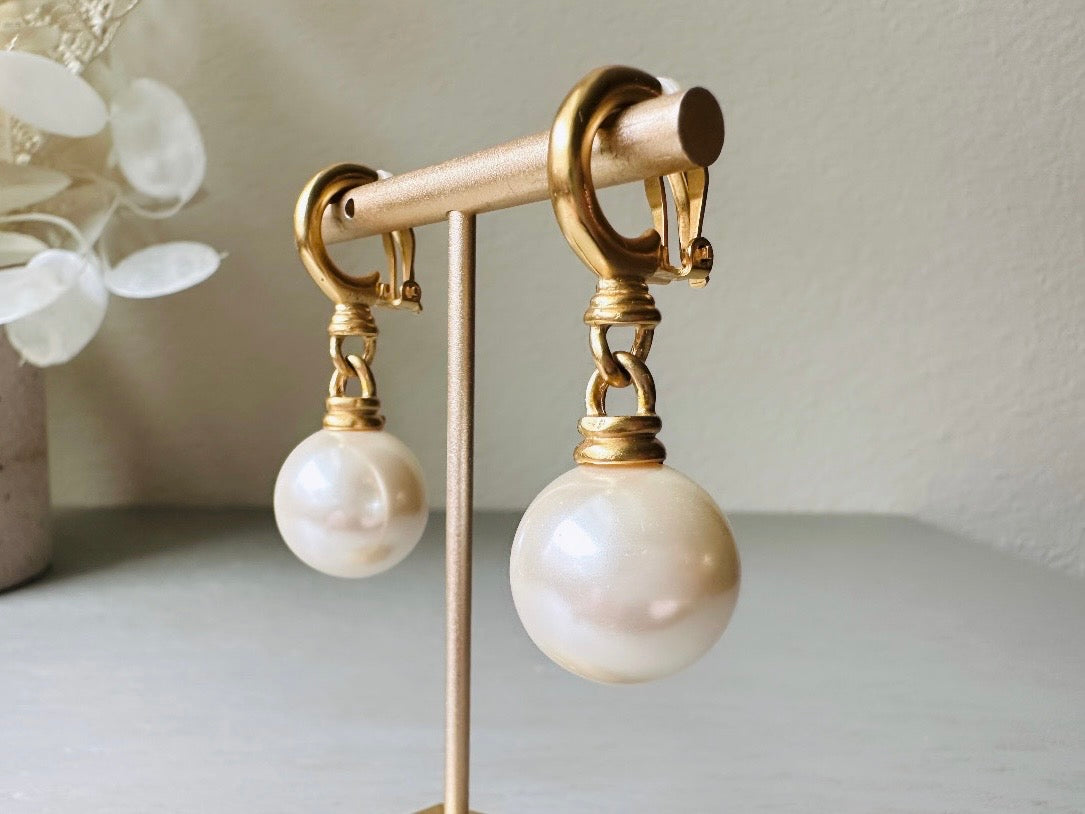 These vintage matte gold and cream pearl drop earrings are clip ons, shown on light gray background with white flowers. They are very elegant. They give the appearance of half huggie hoops in a beautiful satin matte finish gold, with a striking oversized cream glass pearl. These Etruscan statement drop earrings are very on trend. These earrings are 2 inches long, unsigned in excellent condition.