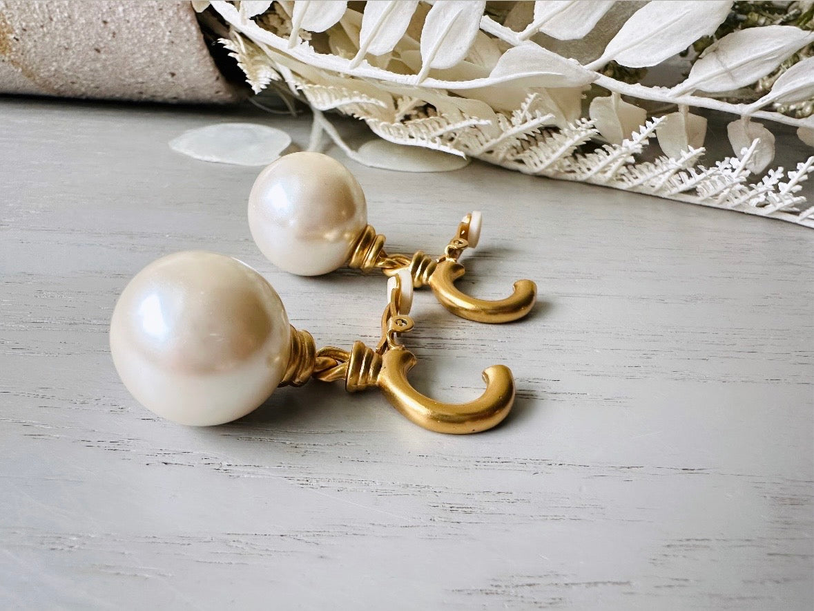 These vintage matte gold and cream pearl drop earrings are clip ons, shown on light gray background with white flowers. They are very elegant. They give the appearance of half huggie hoops in a beautiful satin matte finish gold, with a striking oversized cream glass pearl. These Etruscan statement drop earrings are very on trend. These earrings are 2 inches long, unsigned in excellent condition.