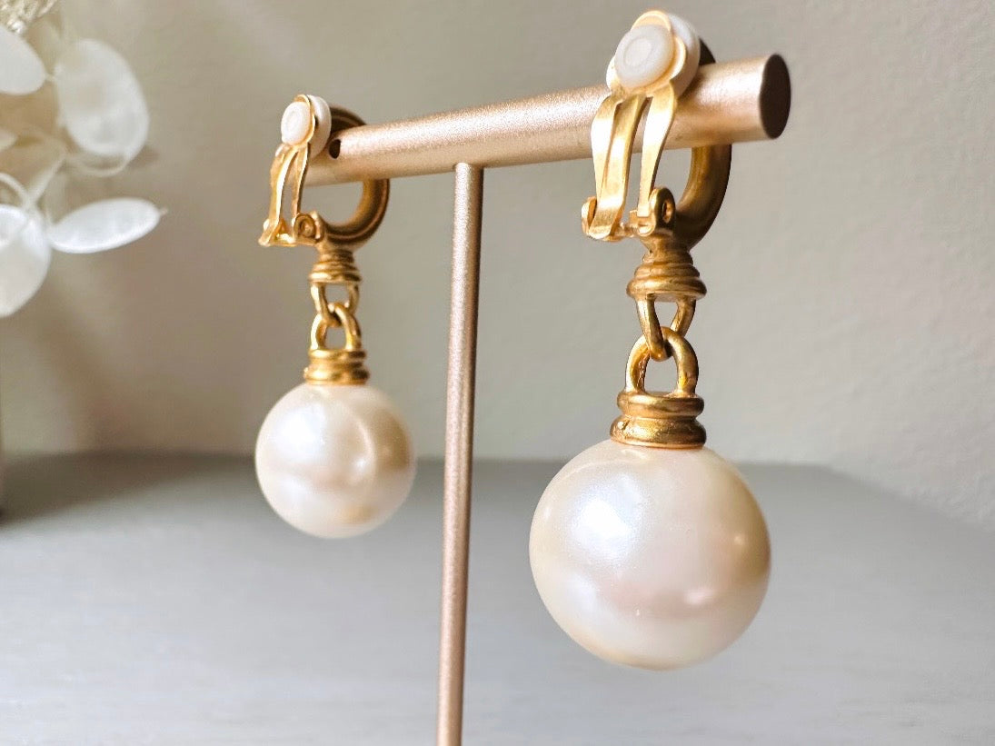 These vintage matte gold and cream pearl drop earrings are clip ons, shown on light gray background with white flowers. They are very elegant. They give the appearance of half huggie hoops in a beautiful satin matte finish gold, with a striking oversized cream glass pearl. These Etruscan statement drop earrings are very on trend. These earrings are 2 inches long, unsigned in excellent condition.