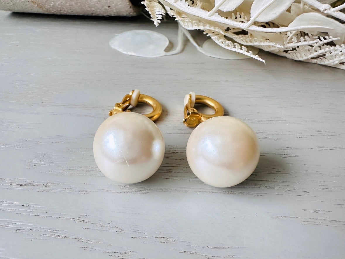 These vintage matte gold and cream pearl drop earrings are clip ons, shown on light gray background with white flowers. They are very elegant. They give the appearance of half huggie hoops in a beautiful satin matte finish gold, with a striking oversized cream glass pearl. These Etruscan statement drop earrings are very on trend. These earrings are 2 inches long, unsigned in excellent condition.