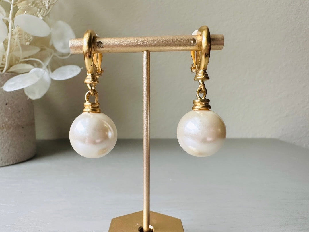 These vintage matte gold and cream pearl drop earrings are clip ons, shown on light gray background with white flowers. They are very elegant. They give the appearance of half huggie hoops in a beautiful satin matte finish gold, with a striking oversized cream glass pearl. These Etruscan statement drop earrings are very on trend. These earrings are 2 inches long, unsigned in excellent condition.