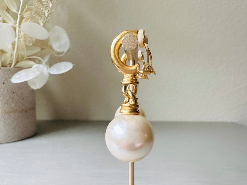 These vintage matte gold and cream pearl drop earrings are clip ons, shown on light gray background with white flowers. They are very elegant. They give the appearance of half huggie hoops in a beautiful satin matte finish gold, with a striking oversized cream glass pearl. These Etruscan statement drop earrings are very on trend. These earrings are 2 inches long, unsigned in excellent condition.