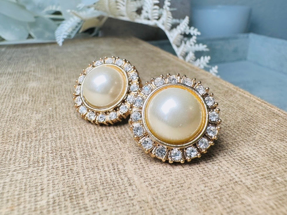 These gold and cream pearl button earrings are a classic old money aesthetic pair from the 80s. Featuring a beautiful cream faux pearl cabochon surrounded by a prong set rhinestone border. These clip on earrings are both unique and a timeless classic, old Hollywood glam style in great vintage condition. 

Dimensions: about 1", please view all pics for a visual size reference.

