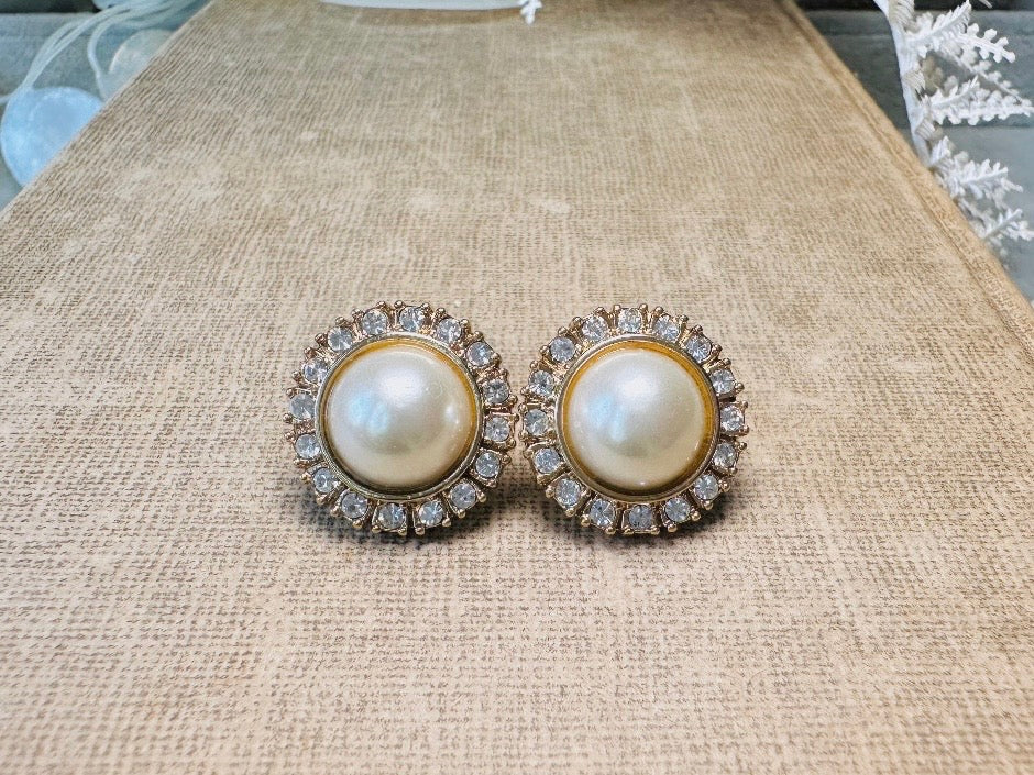 These gold and cream pearl button earrings are a classic old money aesthetic pair from the 80s. Featuring a beautiful cream faux pearl cabochon surrounded by a prong set rhinestone border. These clip on earrings are both unique and a timeless classic, old Hollywood glam style in great vintage condition. 

Dimensions: about 1", please view all pics for a visual size reference.

