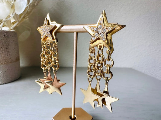 A pair of vintage gold star dangle earrings with a large gold star base with another layered star that is covered in pave set rhinestones. From this star three chain tassels drop, each with a gold star charm dangling from the end. These are clip on style, and in great condition. A fantastic 1980s pair!
