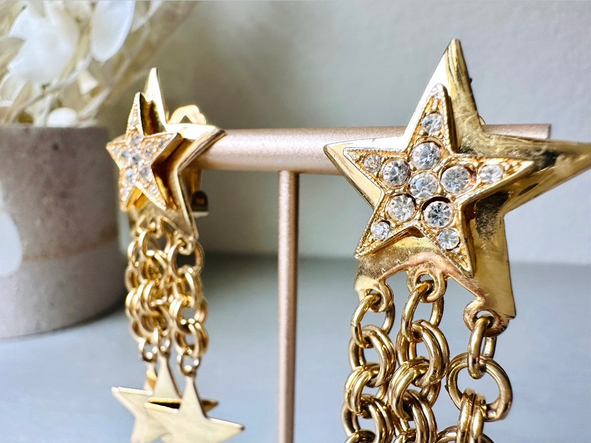 A pair of vintage gold star dangle earrings with a large gold star base with another layered star that is covered in pave set rhinestones. From this star three chain tassels drop, each with a gold star charm dangling from the end. These are clip on style, and in great condition. A fantastic 1980s pair!

