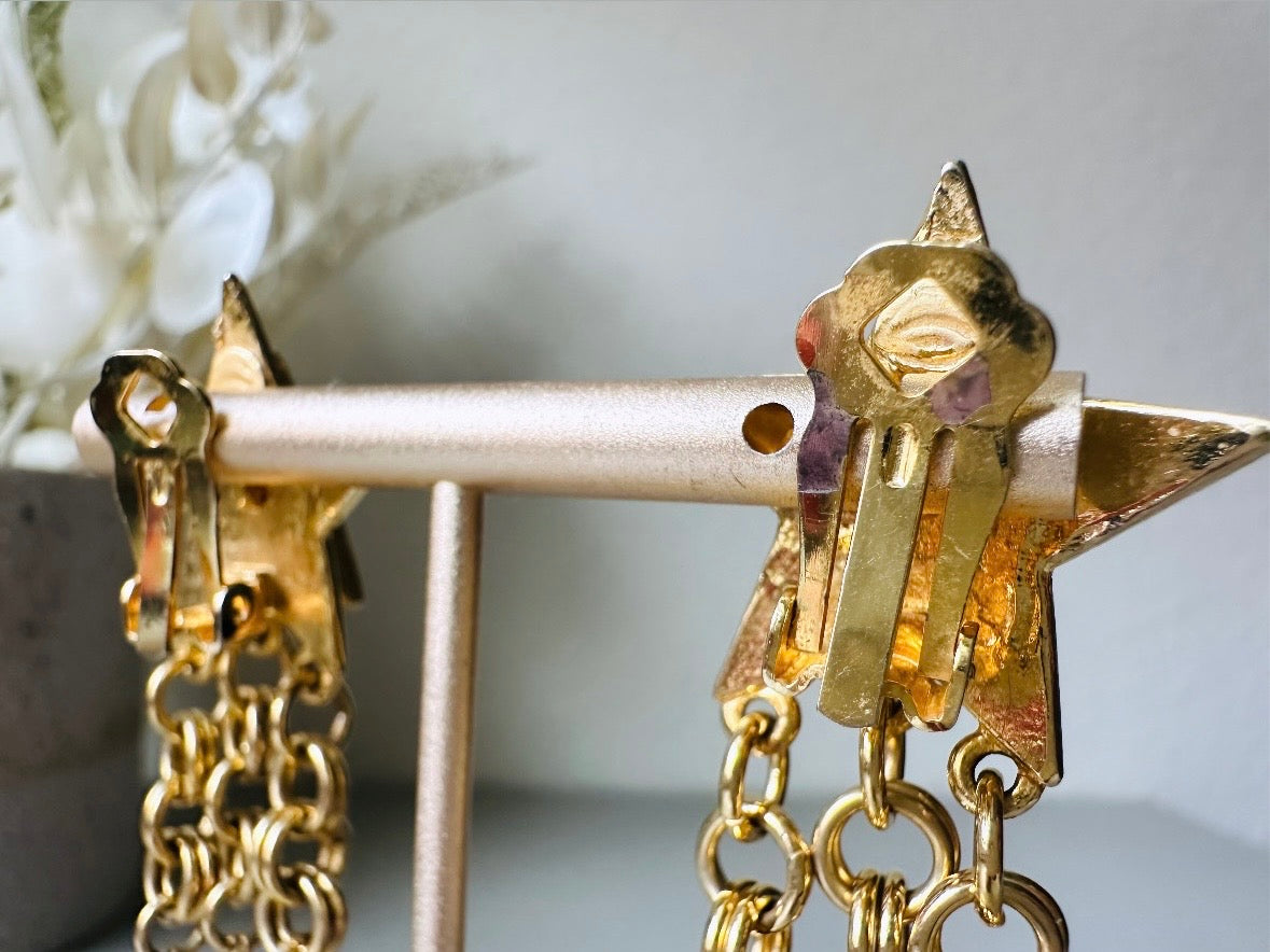 A pair of vintage gold star dangle earrings with a large gold star base with another layered star that is covered in pave set rhinestones. From this star three chain tassels drop, each with a gold star charm dangling from the end. These are clip on style, and in great condition. A fantastic 1980s pair!

