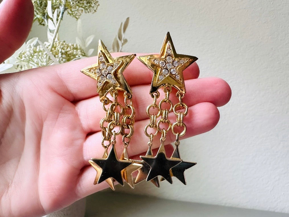 A pair of vintage gold star dangle earrings with a large gold star base with another layered star that is covered in pave set rhinestones. From this star three chain tassels drop, each with a gold star charm dangling from the end. These are clip on style, and in great condition. A fantastic 1980s pair!
Held in hand for size reference.
