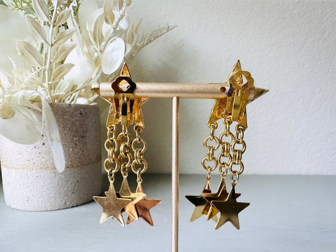 A pair of vintage gold star dangle earrings with a large gold star base with another layered star that is covered in pave set rhinestones. From this star three chain tassels drop, each with a gold star charm dangling from the end. These are clip on style, and in great condition. A fantastic 1980s pair!
