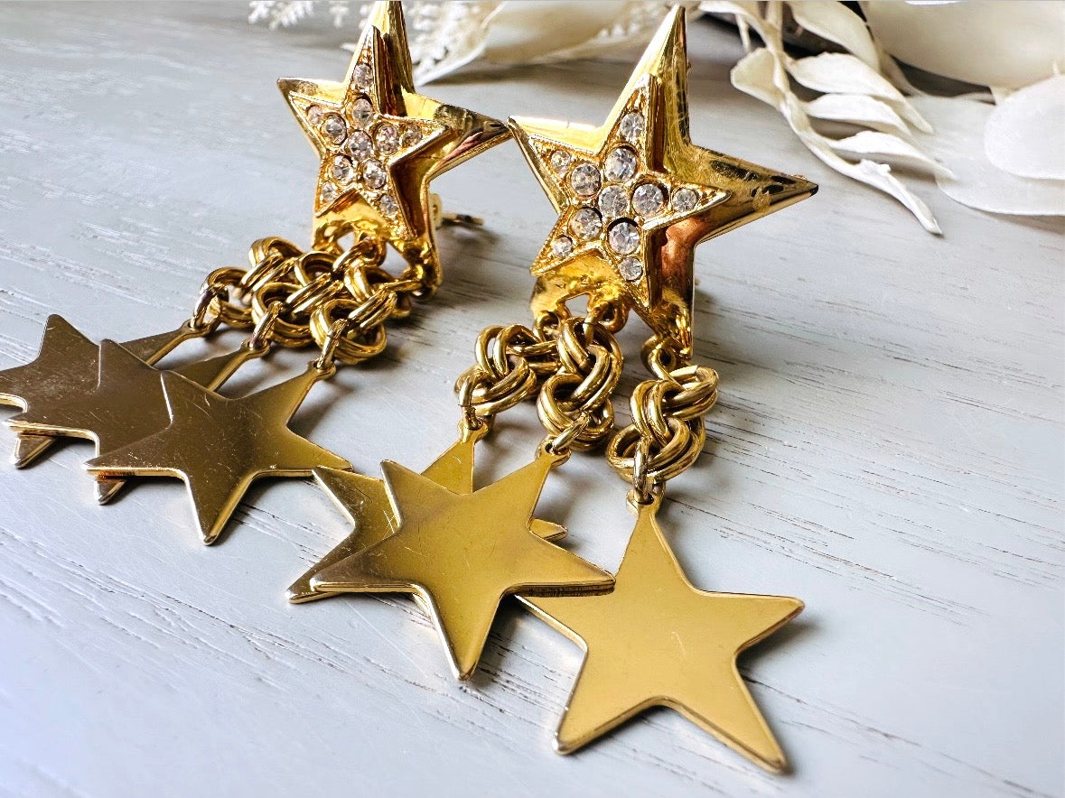A pair of vintage gold star dangle earrings with a large gold star base with another layered star that is covered in pave set rhinestones. From this star three chain tassels drop, each with a gold star charm dangling from the end. These are clip on style, and in great condition. A fantastic 1980s pair!
