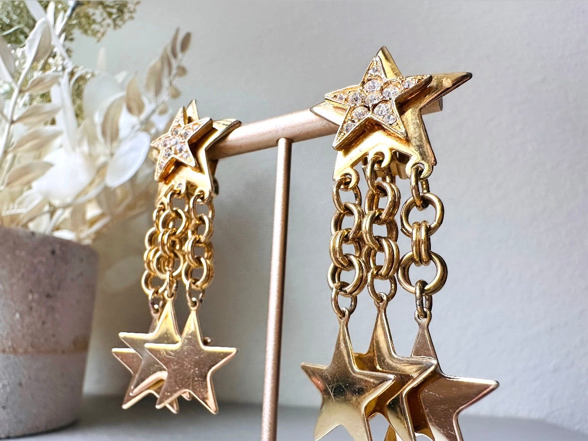 A pair of vintage gold star dangle earrings with a large gold star base with another layered star that is covered in pave set rhinestones. From this star three chain tassels drop, each with a gold star charm dangling from the end. These are clip on style, and in great condition. A fantastic 1980s pair!
