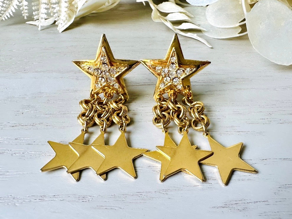 A pair of vintage gold star dangle earrings with a large gold star base with another layered star that is covered in pave set rhinestones. From this star three chain tassels drop, each with a gold star charm dangling from the end. These are clip on style, and in great condition. A fantastic 1980s pair!
