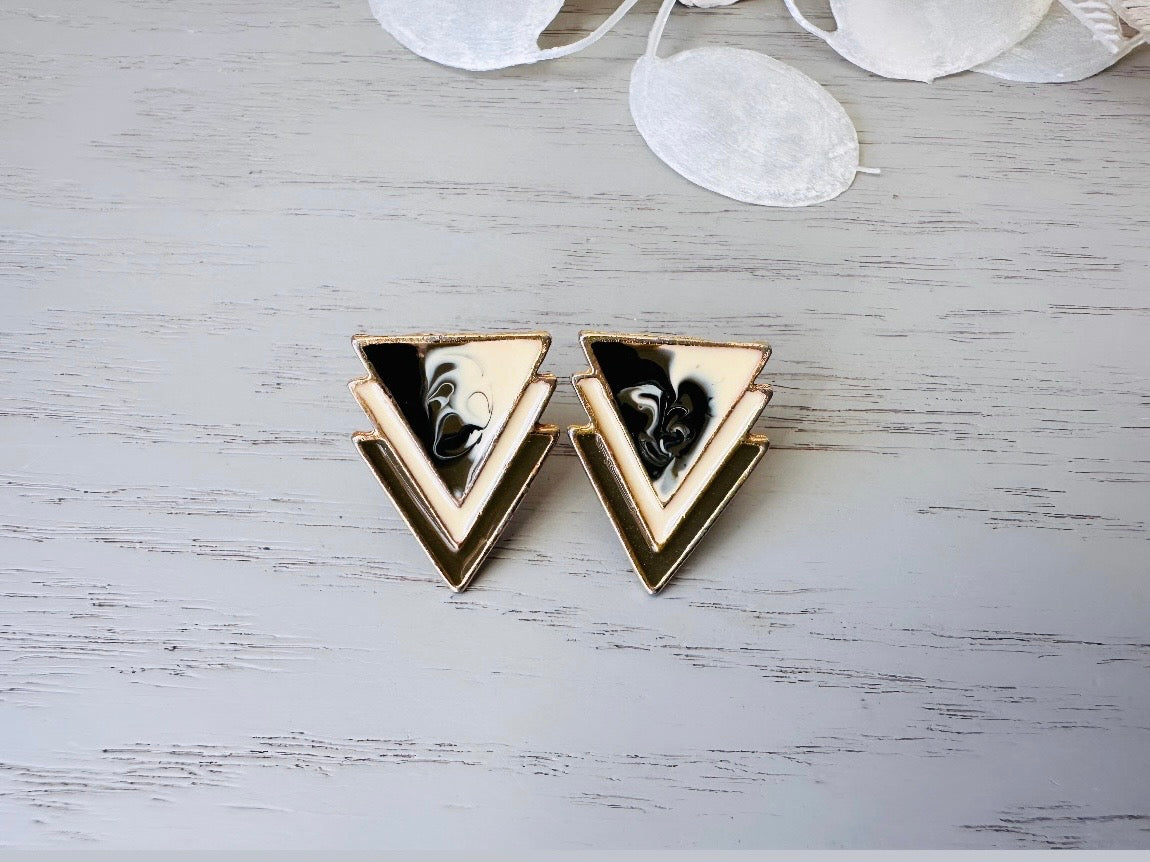 A pair of black, cream, and gold vintage triangle shaped earrings from the 1980s. The earrings are made of layered triangles with a marbled design.