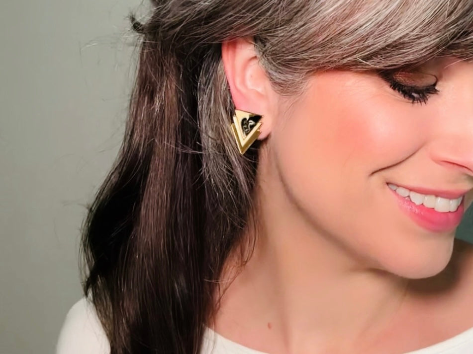 A pair of black, cream, and gold vintage triangle shaped earrings from the 1980s. The earrings are made of layered triangles with a marbled design. Modelled for size reference.