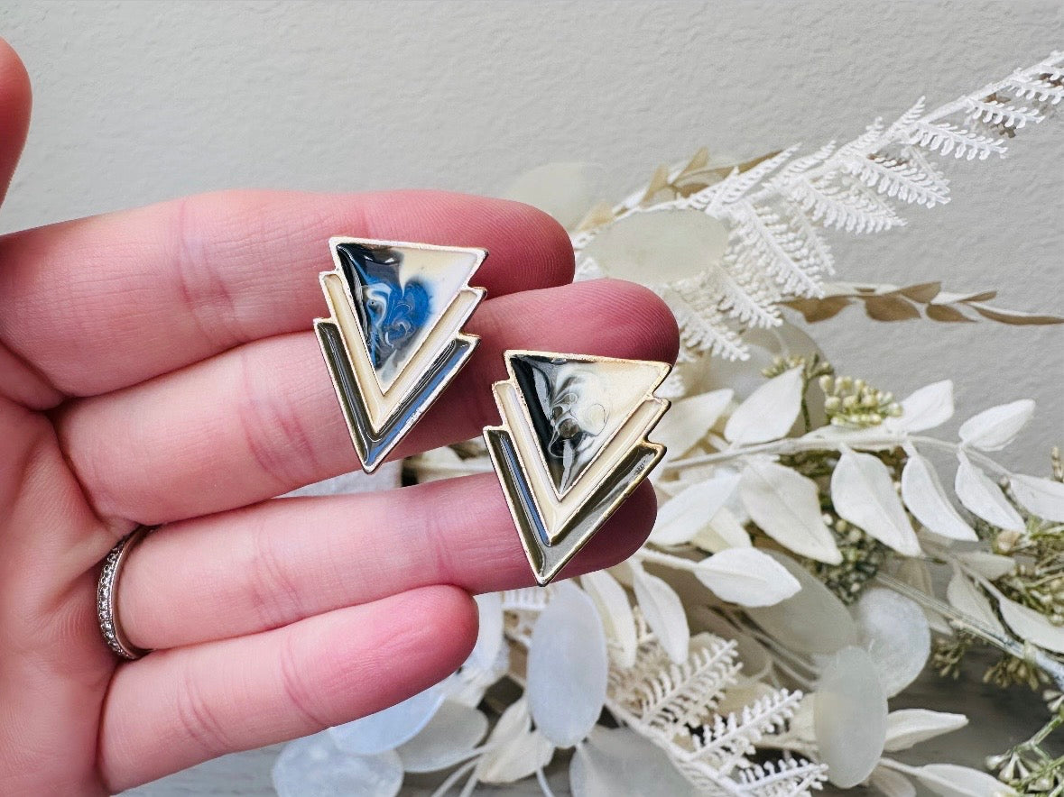 A pair of black, cream, and gold vintage triangle shaped earrings from the 1980s. The earrings are made of layered triangles with a marbled design. Held in hand for size reference.