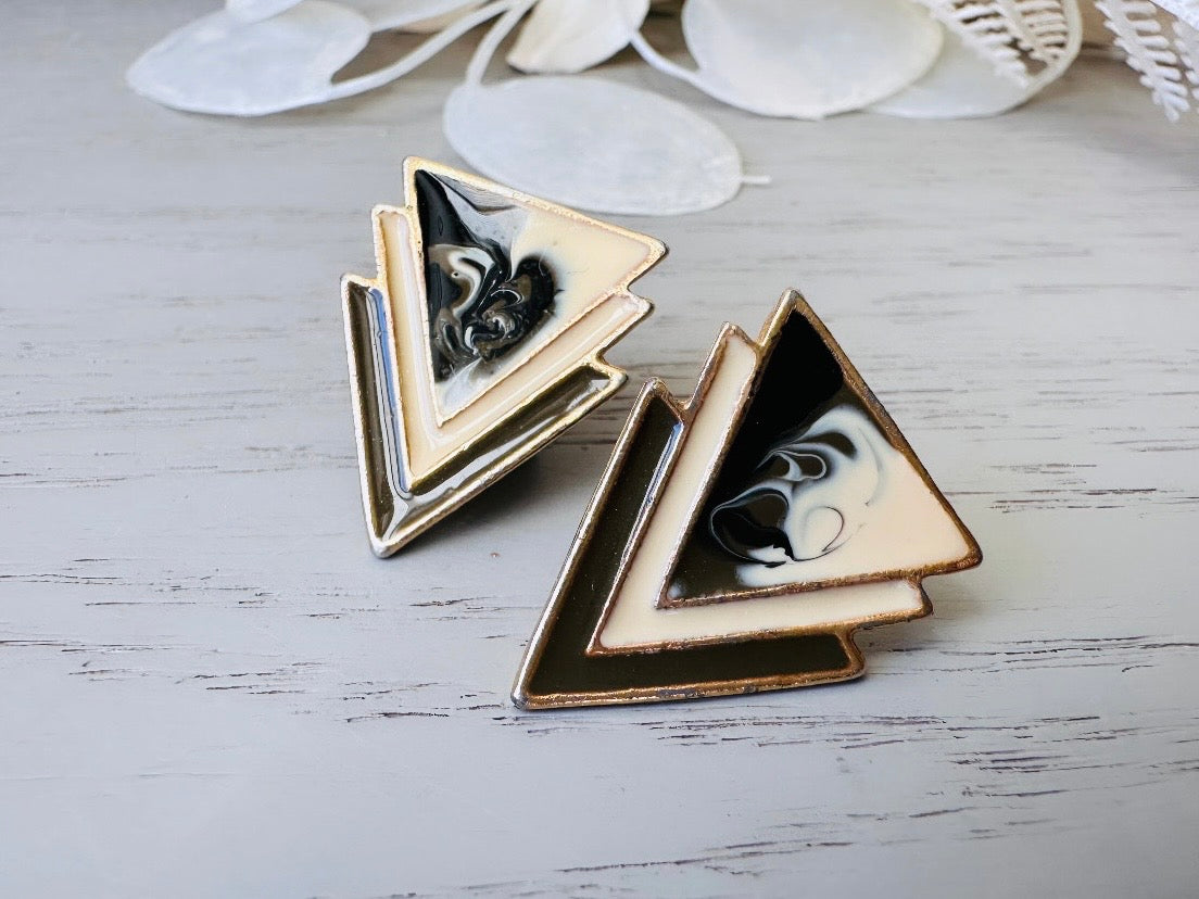 A pair of black, cream, and gold vintage triangle shaped earrings from the 1980s. The earrings are made of layered triangles with a marbled design.