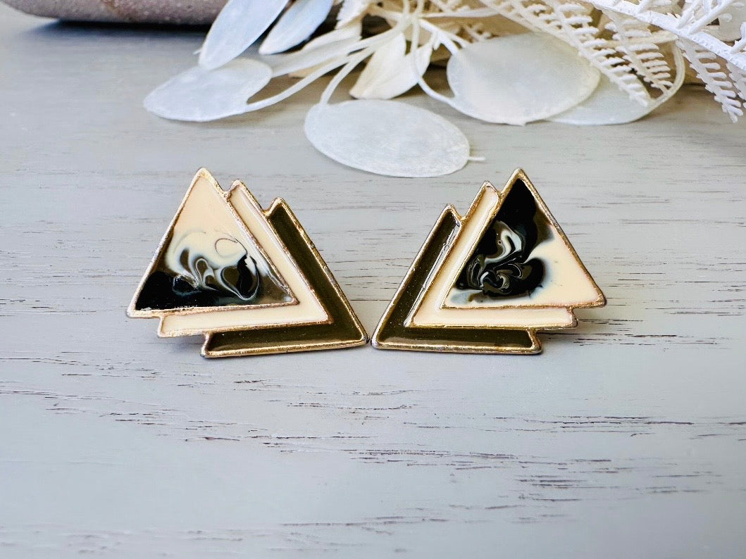 A pair of black, cream, and gold vintage triangle shaped earrings from the 1980s. The earrings are made of layered triangles with a marbled design.