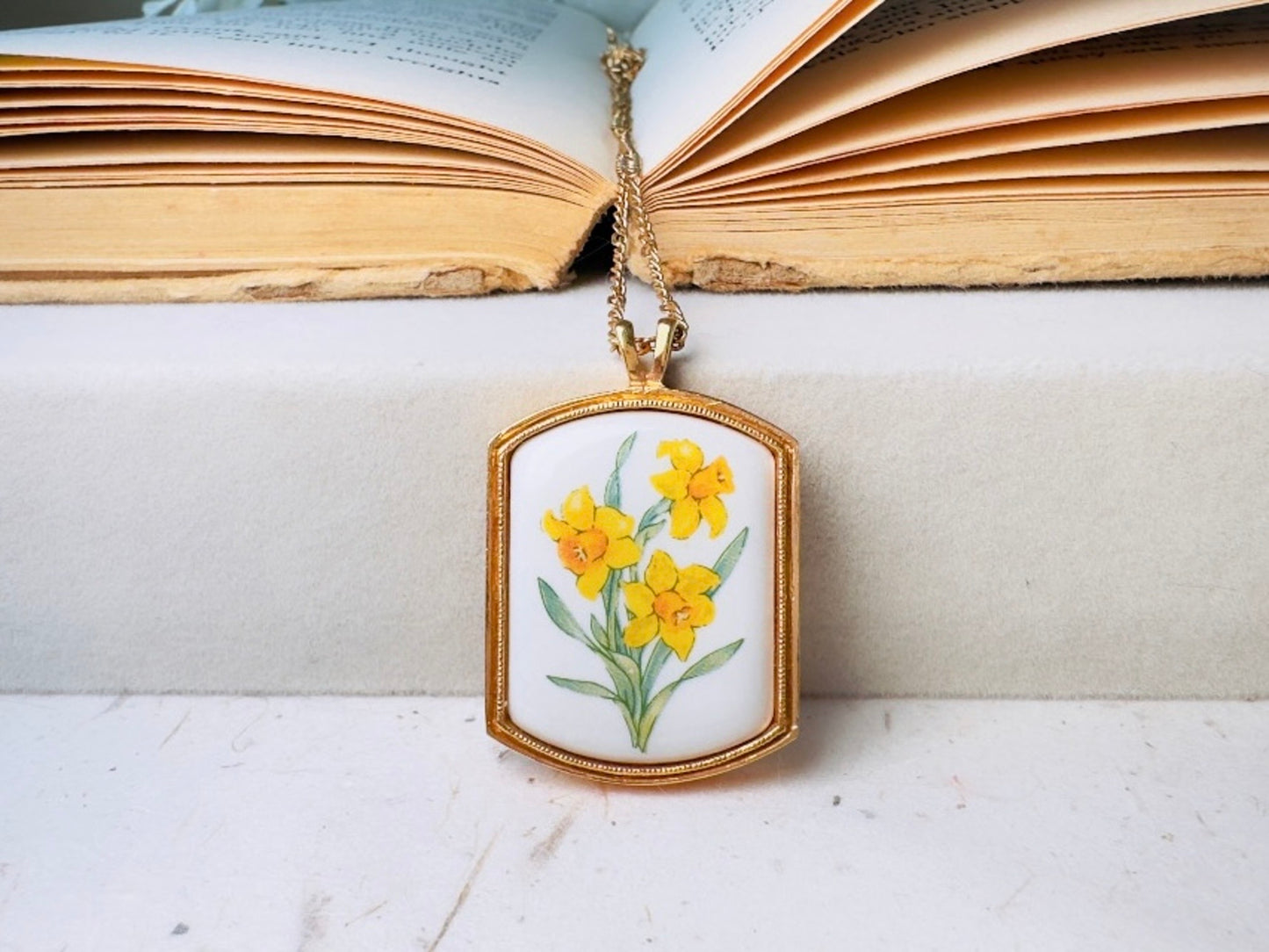 A gold-tone pendant necklace featuring a white enamel background with a painted yellow daffodil flower design, hanging from a gold chain in front of an open vintage book.