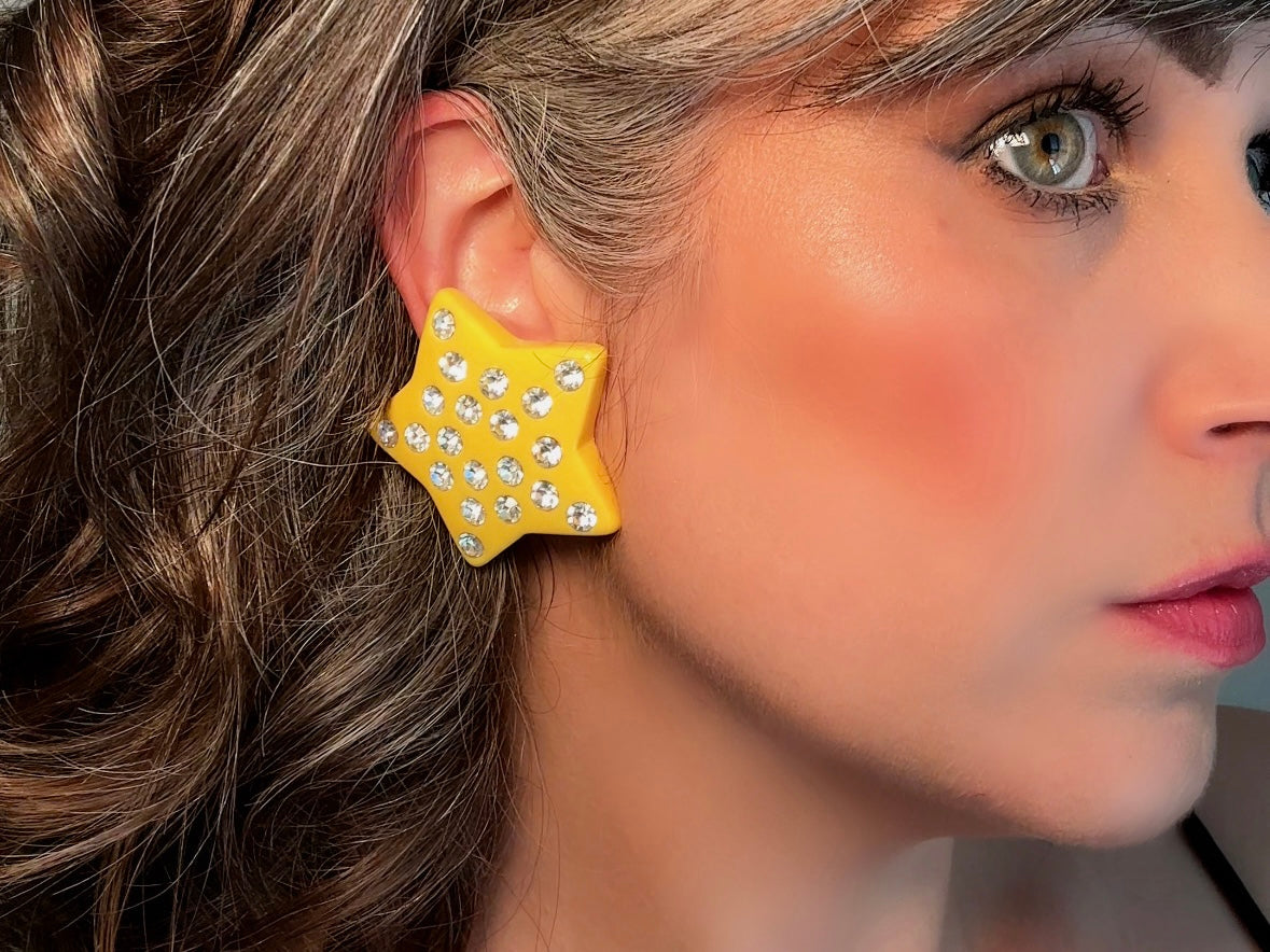 A pair of yellow star earrings from Piggle and Pop. A highly collectible pair of 1980s statement earrings. These yellow star earrings are bright banana yellow opaque lucite with diamond rhinestone accents. Huge vintage star clip ons with major rockstar vibes. on model Bethan Jayne.