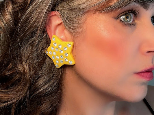 A pair of yellow star earrings from Piggle and Pop. A highly collectible pair of 1980s statement earrings. These yellow star earrings are bright banana yellow opaque lucite with diamond rhinestone accents. Huge vintage star clip ons with major rockstar vibes. on model Bethan Jayne.