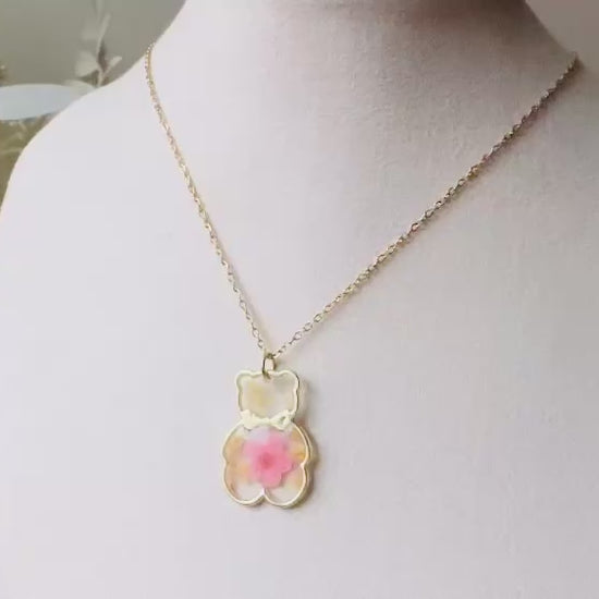 An adorable handmade teddy bear necklace. The necklace is made of 18kt gold and it has a cute teddy bear charm. The charm is clear with pink and yellow real pressed flowers. The bear is trimmed in gold and has a little gold bowtie.