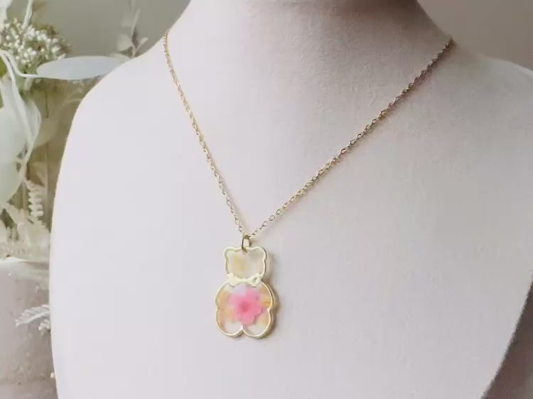 An adorable handmade teddy bear necklace. The necklace is made of 18kt gold and it has a cute teddy bear charm. The charm is clear with pink and yellow real pressed flowers. The bear is trimmed in gold and has a little gold bowtie.