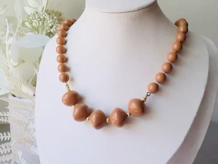 A vintage Park Lane designer necklace. The necklace is composed of 6mm caramel round beads with 5 bigger rondelle beads in the center with tiny gold spacer beads between. The necklace is on a bust display on a light gray wood table with a floral background. This is a video to show the necklace.