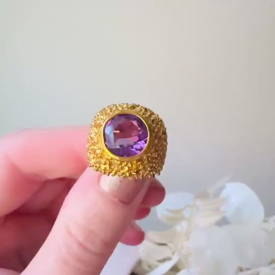 A vintage 1970s cocktail ring, that is gold and purple, The gold band has a raised textured gold nugget surface with a beautiful faceted round cut vibrant amethyst purple in the center. Video to show all angles.
