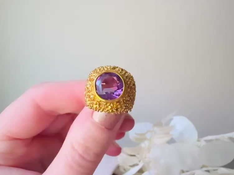 A vintage 1970s cocktail ring, that is gold and purple, The gold band has a raised textured gold nugget surface with a beautiful faceted round cut vibrant amethyst purple in the center. Video to show all angles.