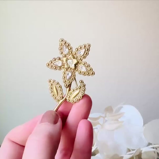 A beautiful vintage flower brooch by the brand 1928. The brooch is a single flower shape that is gold, with textured dotting all around each petal and leaf. There are diamond crystal rhinestones in a teardrop shape in the center of each flower petal and a round crystal in the center. The stem is a smooth shiny gold. The brooch is on the signature background of the shop Piggle and Pop, a light gray wood table with white floral background. This video shows the pin held in a hand for size reference.