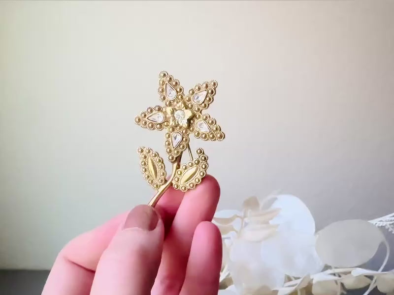 A beautiful vintage flower brooch by the brand 1928. The brooch is a single flower shape that is gold, with textured dotting all around each petal and leaf. There are diamond crystal rhinestones in a teardrop shape in the center of each flower petal and a round crystal in the center. The stem is a smooth shiny gold. The brooch is on the signature background of the shop Piggle and Pop, a light gray wood table with white floral background. This video shows the pin held in a hand for size reference.