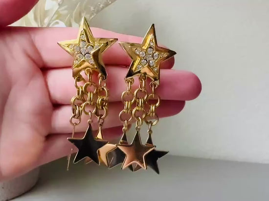 Video of a pair of vintage gold star dangle earrings with a large gold star base with another layered star that is covered in pave set rhinestones. From this star three chain tassels drop, each with a gold star charm dangling from the end. These are clip on style, and in great condition. A fantastic 1980s pair!
