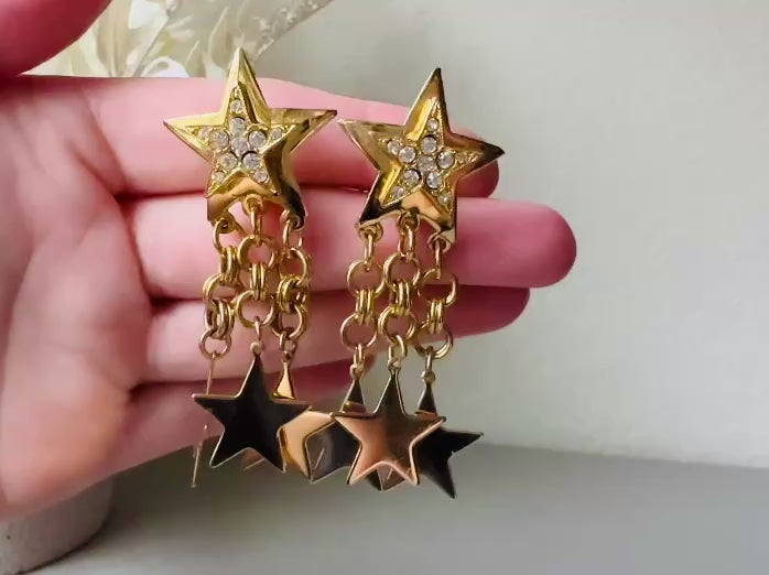 Video of a pair of vintage gold star dangle earrings with a large gold star base with another layered star that is covered in pave set rhinestones. From this star three chain tassels drop, each with a gold star charm dangling from the end. These are clip on style, and in great condition. A fantastic 1980s pair!
